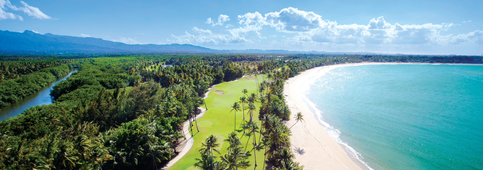 Blick über die Küste Puerto Ricos mit weißem Strand, Palmen, Golfplatz und grünen Bergen im Hintergrund